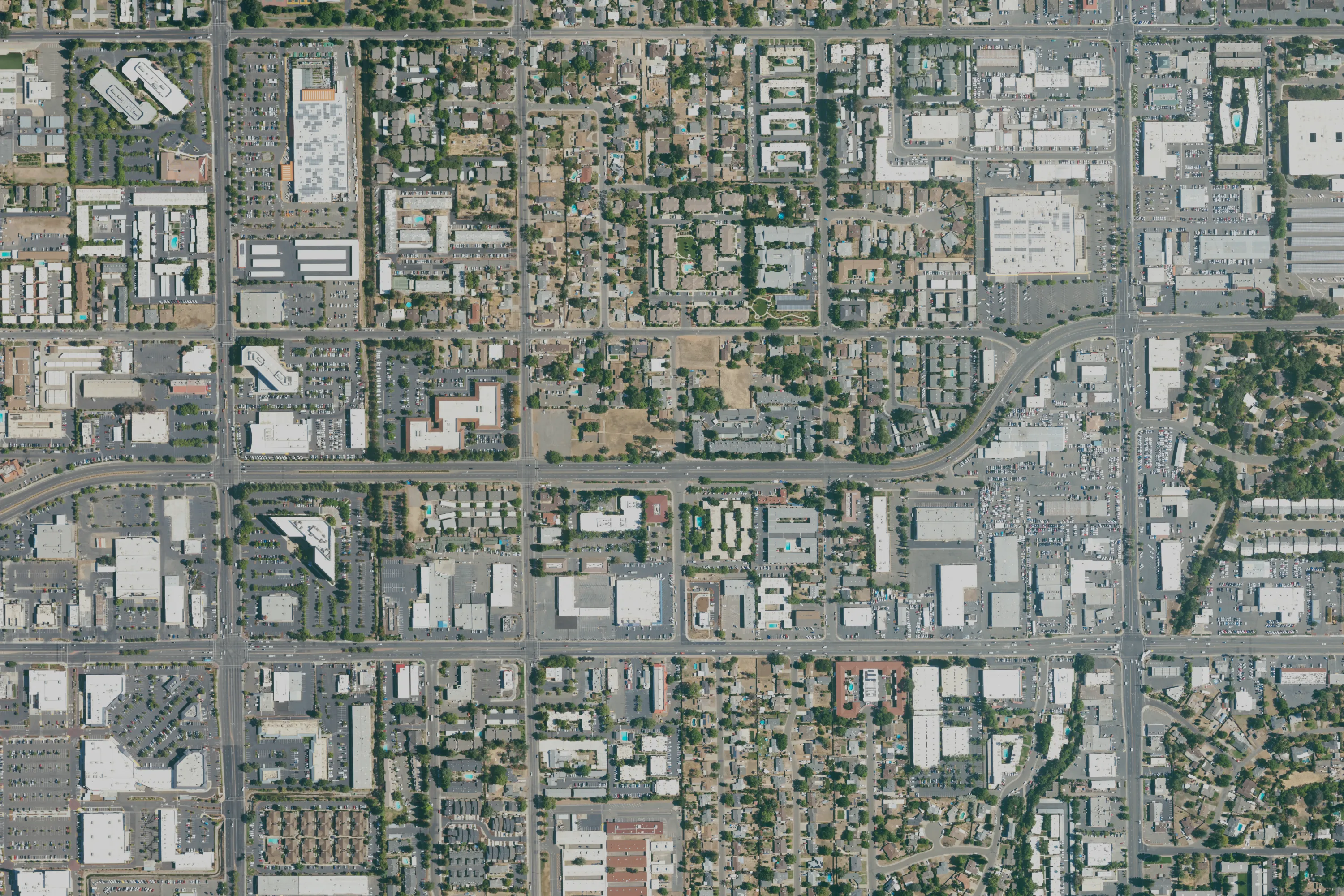 A NAIP aerial photograph of a Sacramento suburb in summer: a grid of arterial roads with low commercial buildings and car parks, blocks of houses and apartment complexes among trees, and a few dry vacant lots.