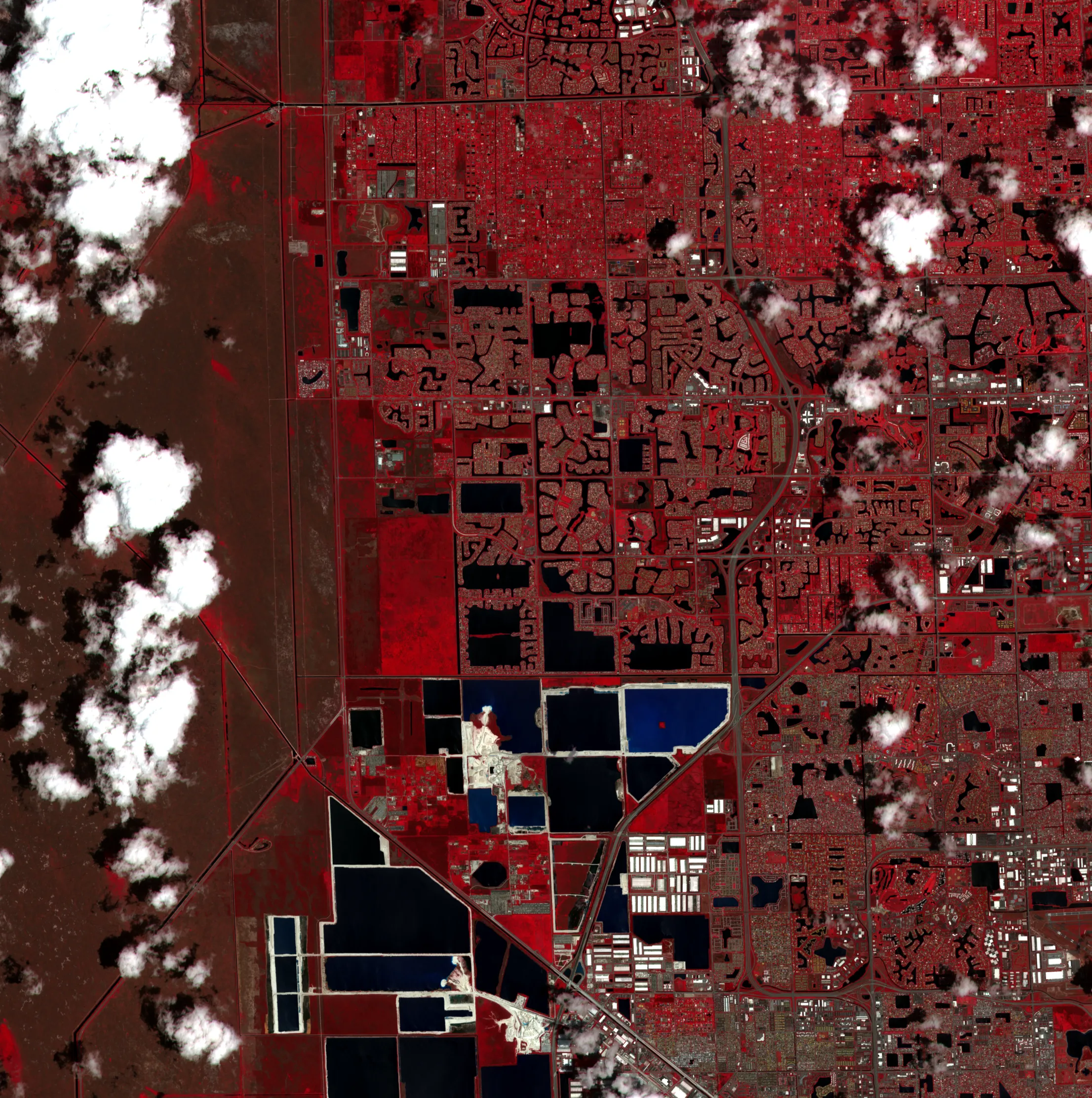 Two Sentinel-2 false-colour views of the same 22-kilometre square of western Broward County, Florida, five weeks apart. Near-infrared is shown as red, so suburban lawns and trees read bright red, water in canals and quarry lakes reads black or deep blue, and the Everglades marsh along the left edge reads dull brown. Scattered cumulus cloud and its shadows sit over both frames, heavier in May.